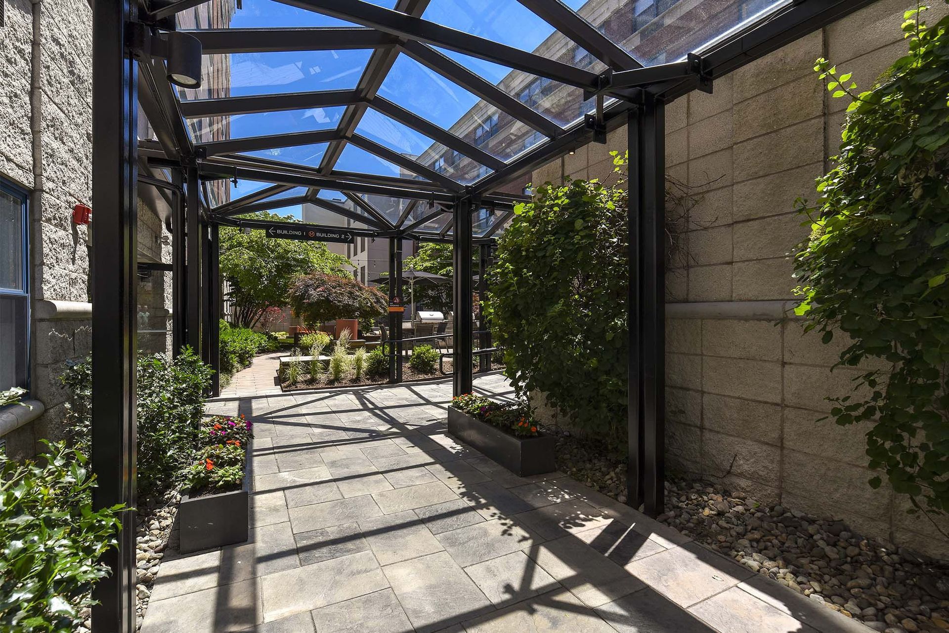 A covered walkway with a glass roof, flanked by brick walls and greenery, leads to a garden with tables and chairs.