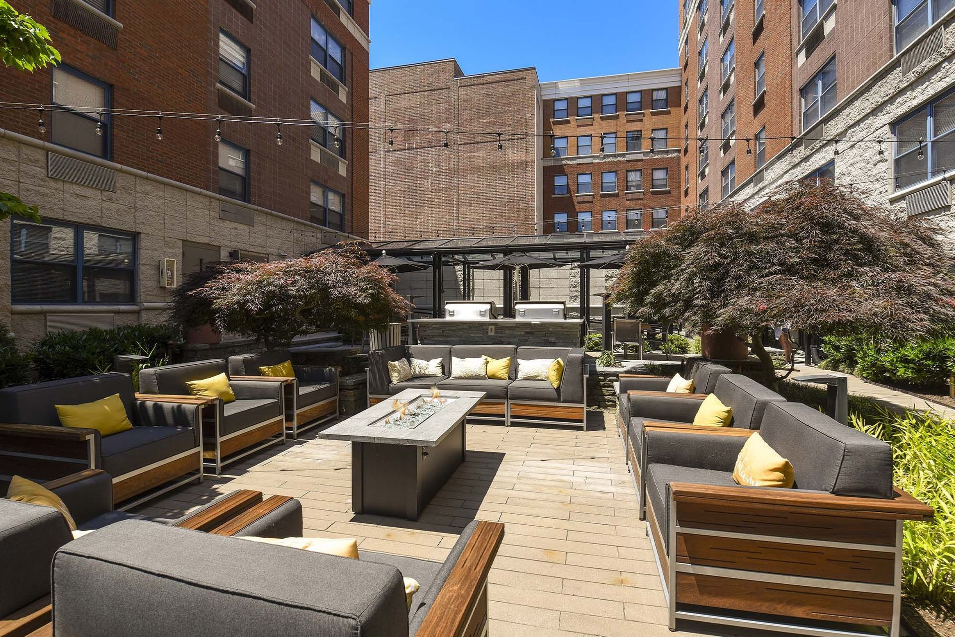 Outdoor patio with gray seating, yellow pillows, a fire pit, and surrounding buildings.