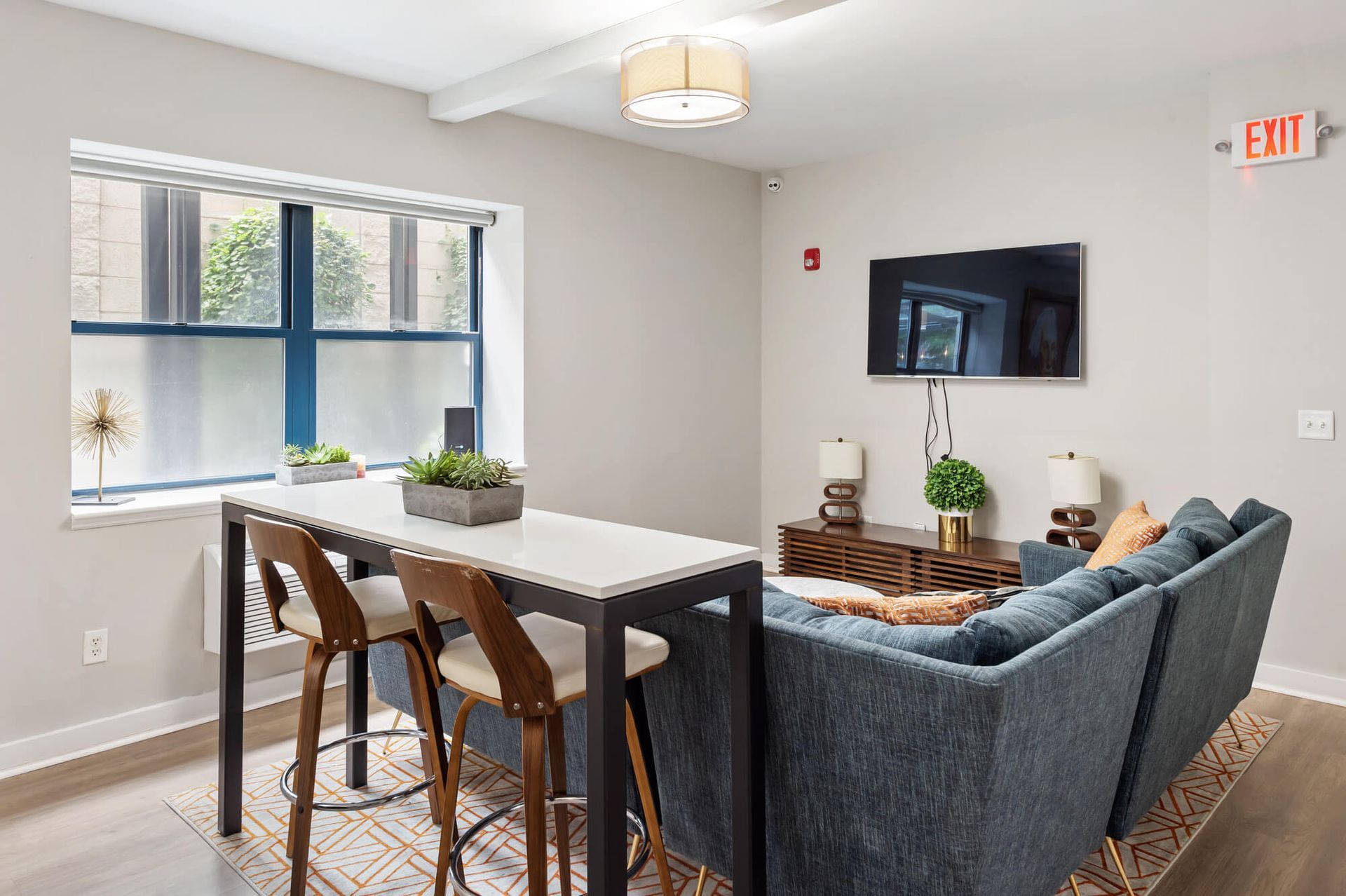 A modern lounge area with a blue sofa, a high table with stools, a TV, and a window.