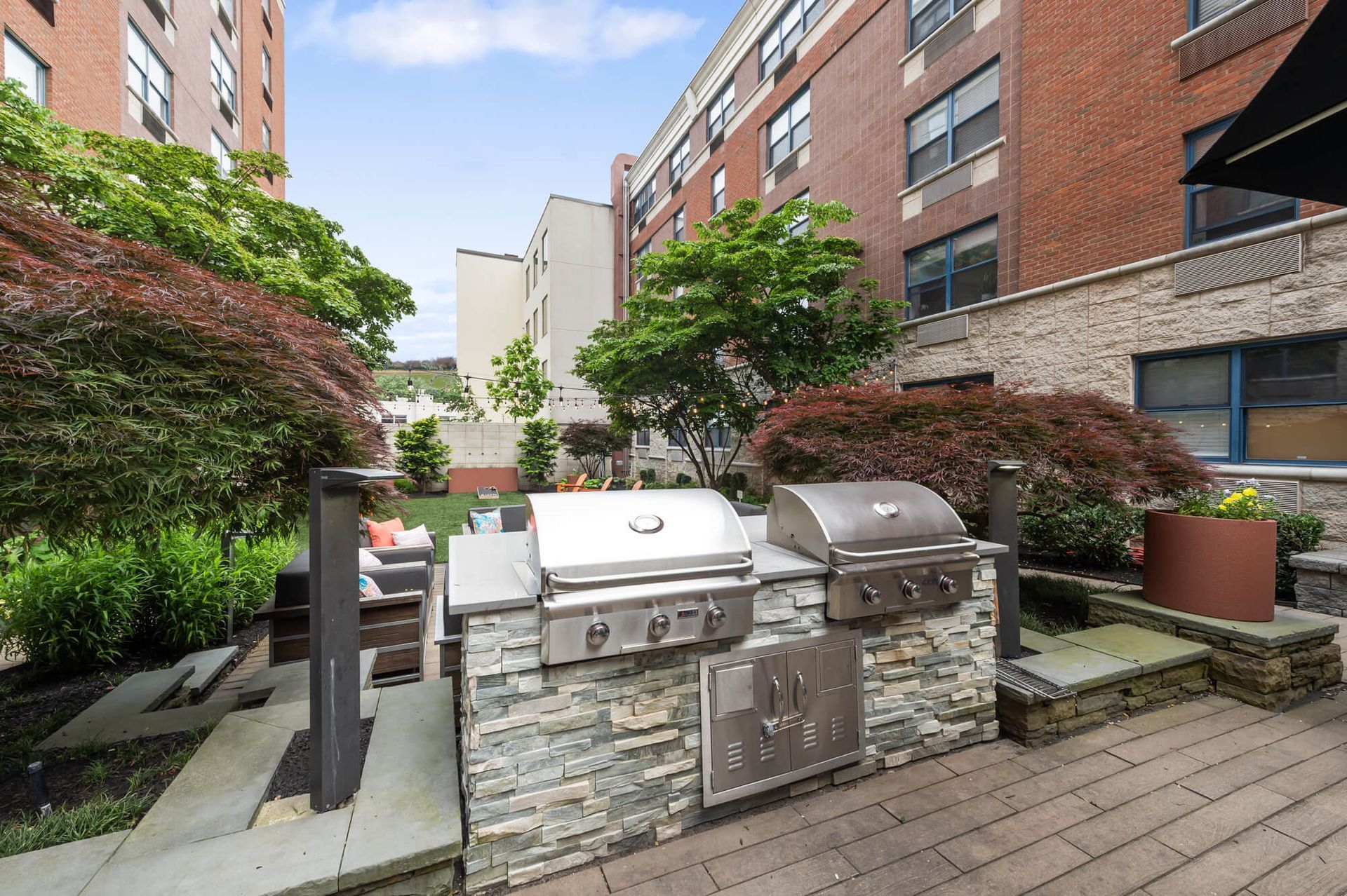 Outdoor kitchen with stone island, stainless steel grills, surrounded by trees and buildings.