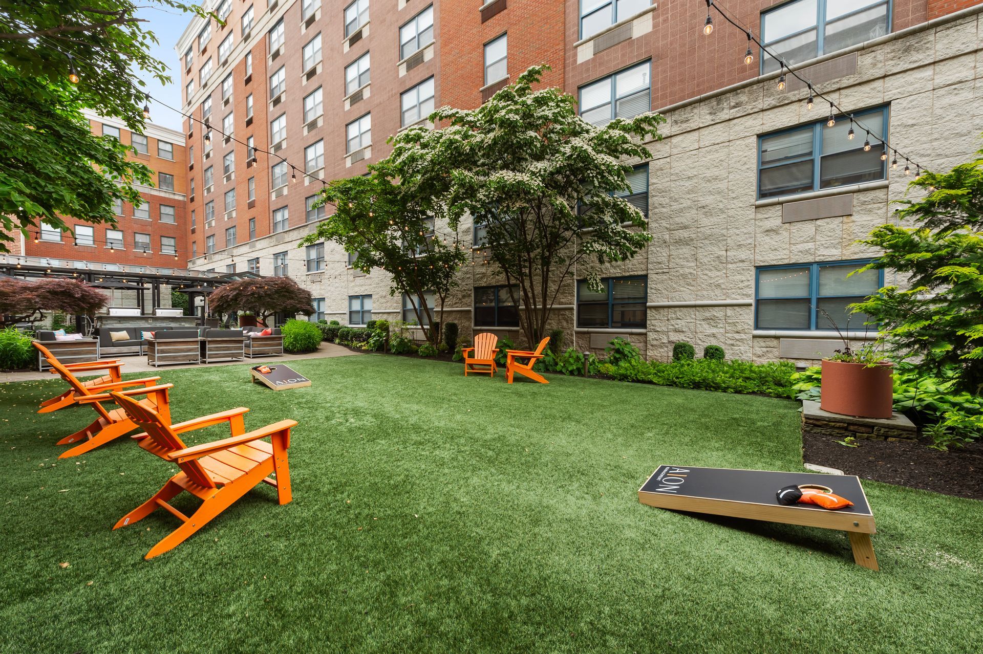 Outdoor lounge area with orange chairs, artificial turf, and a cornhole game next to a brick building.