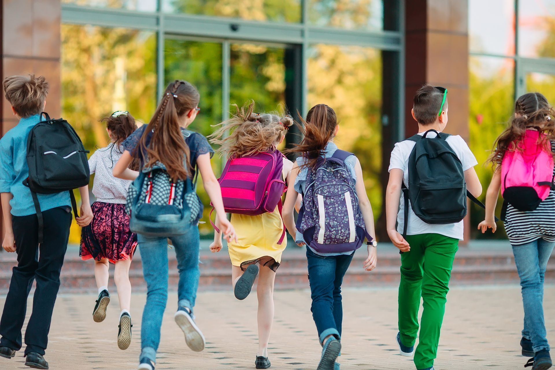 Children with backpacks run toward a school building with glass doors.