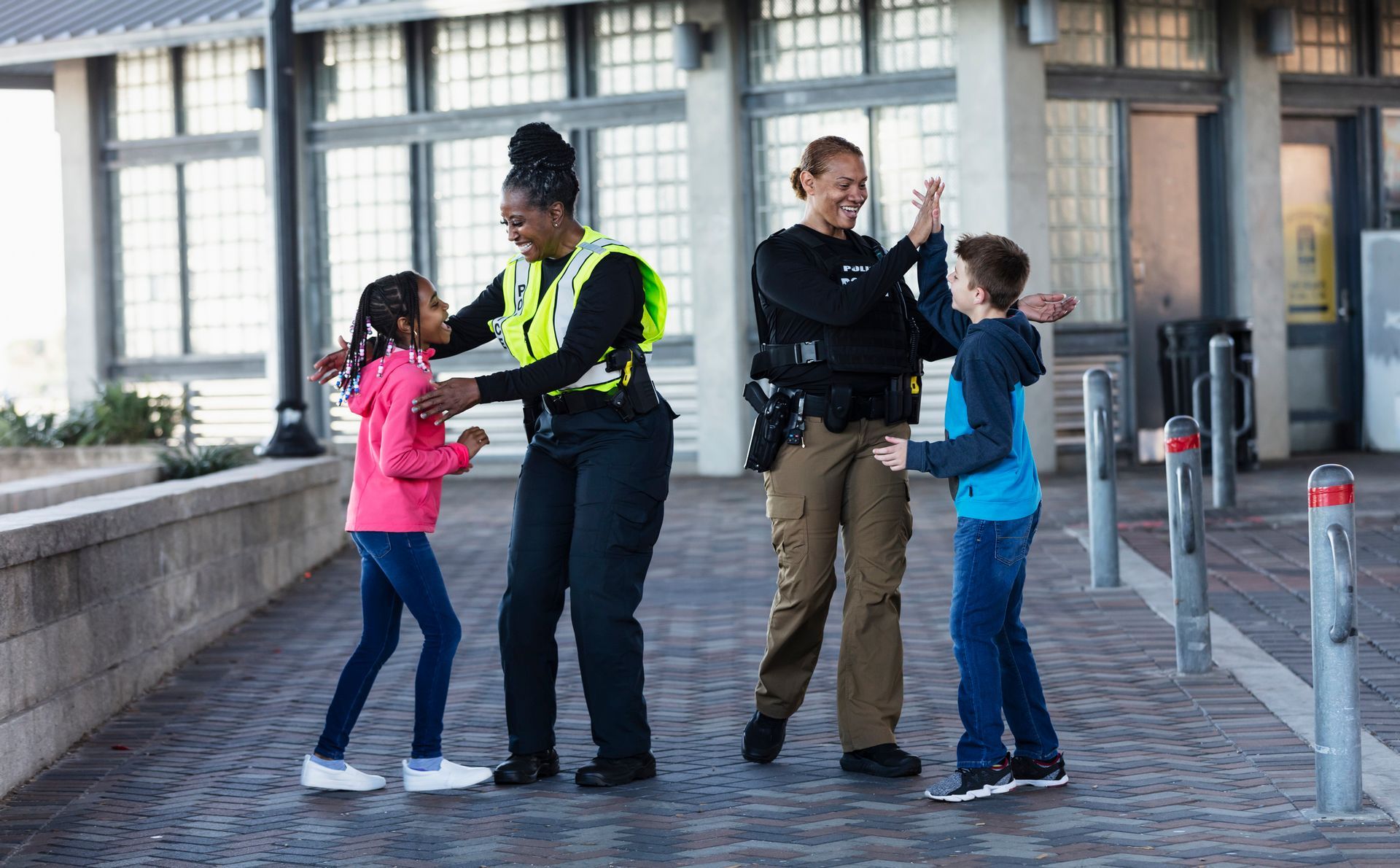 Two police officers high-fiving children outside a building. One officer wears a reflective vest.