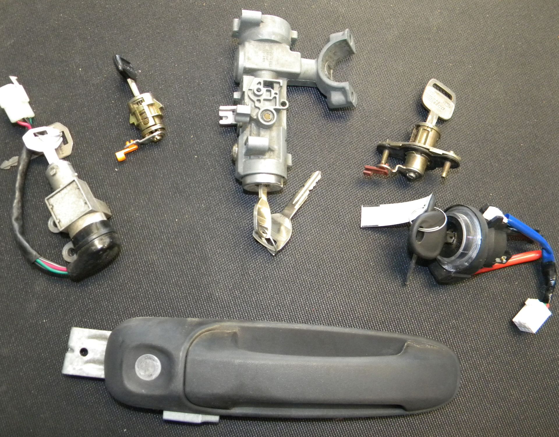 Assorted automotive lock components including ignition cylinders, door lock parts, wiring, and a car door handle displayed on a work surface.