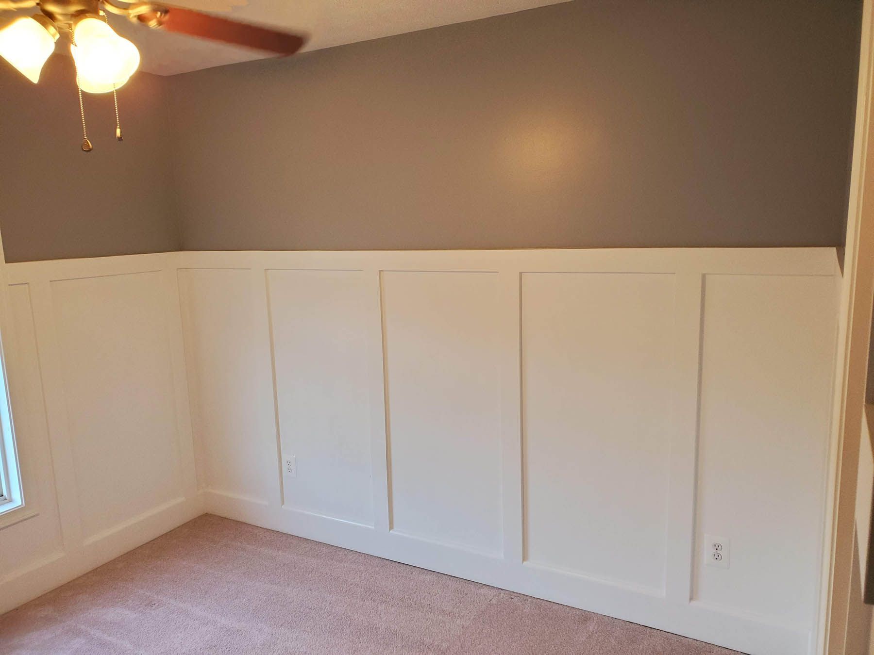White wainscoting on a wall with gray above, beige carpet and a ceiling fan in the corner.