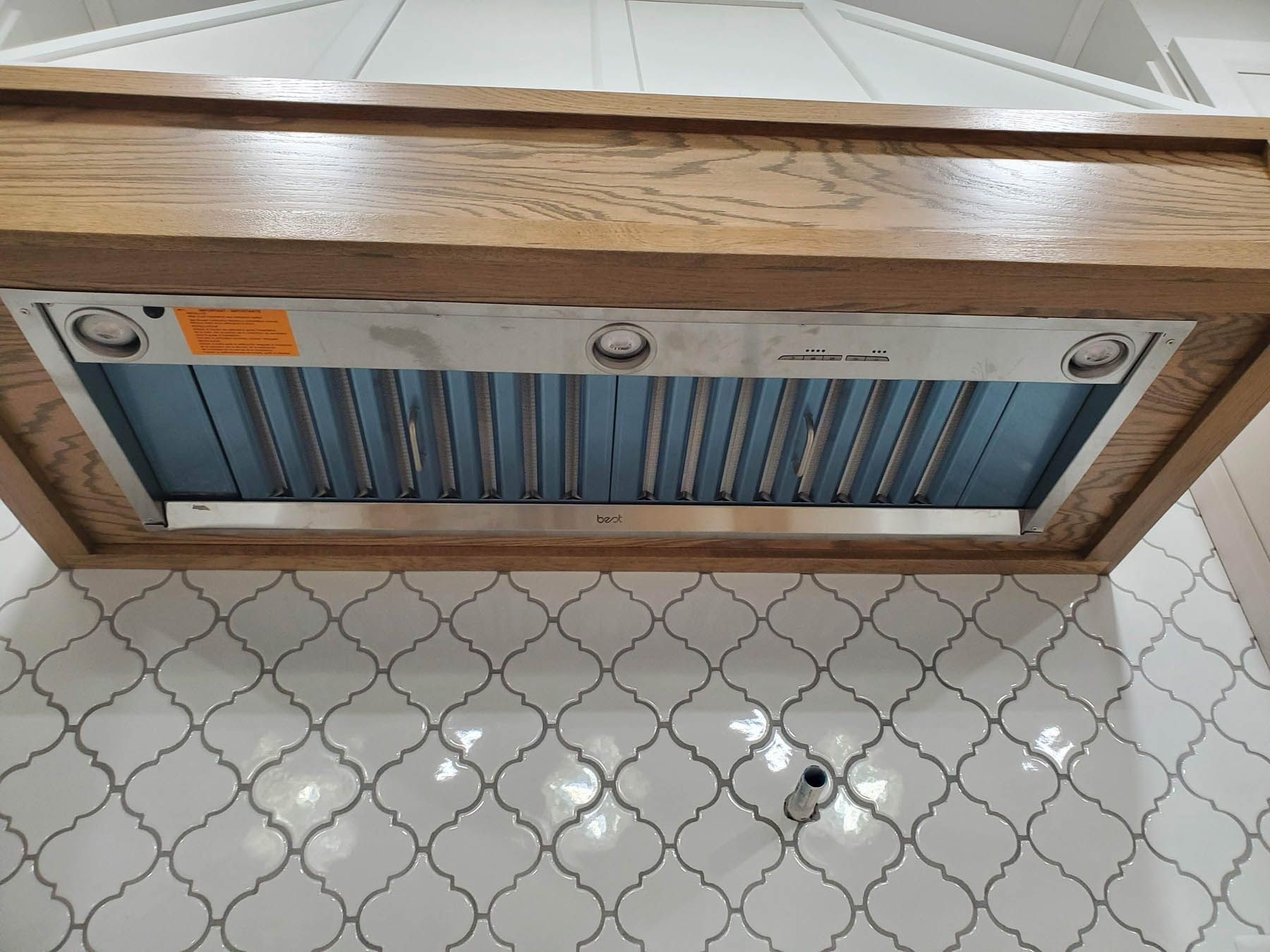 Wooden range hood with stainless steel interior and blue filters over white tile backsplash.