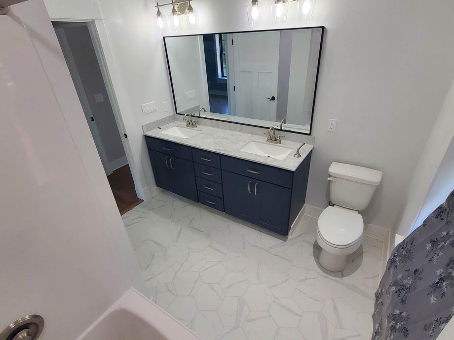 Newly renovated bathroom: navy vanity, double sinks, large mirrors, white hexagon tile floor, toilet, and tub.