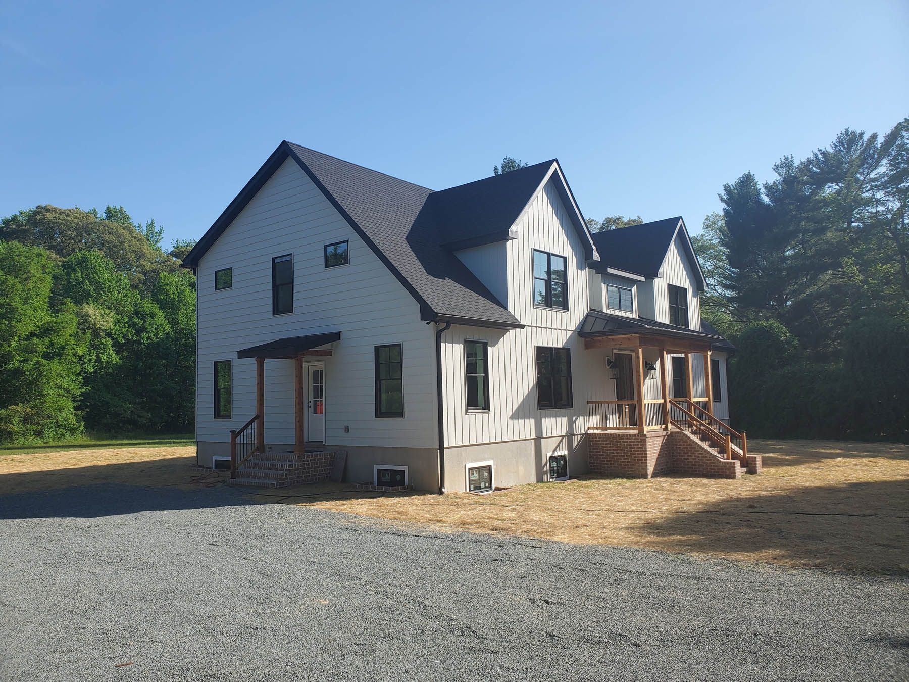 Two-story white farmhouse with black roof and accents; gravel driveway, surrounded by trees, sunny day.