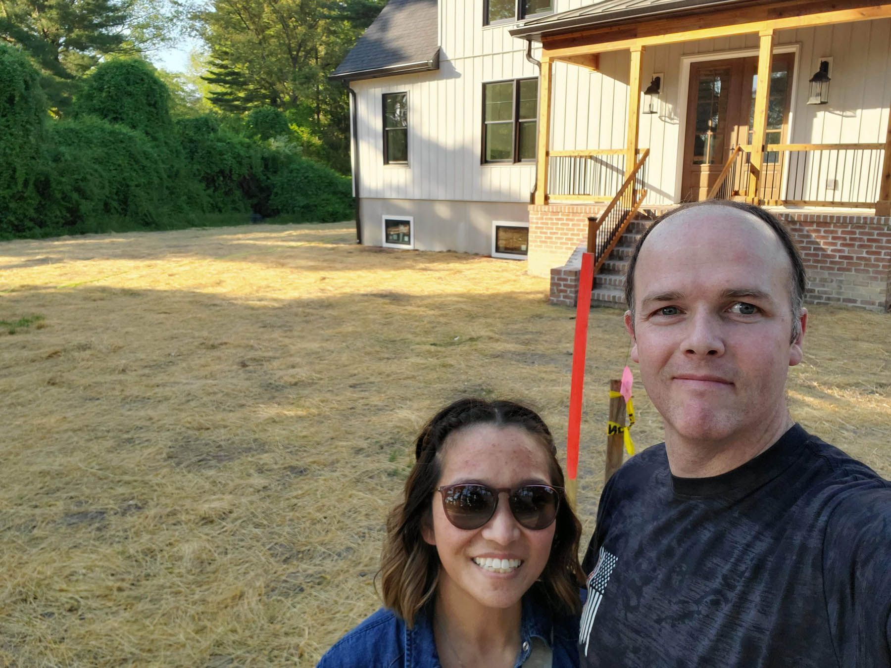 Couple smiling in front of new house with bare yard; sunlight; green foliage.