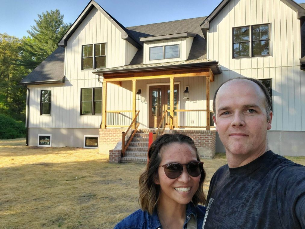 Couple smiling in front of a newly built farmhouse-style house with a porch and a brown door.