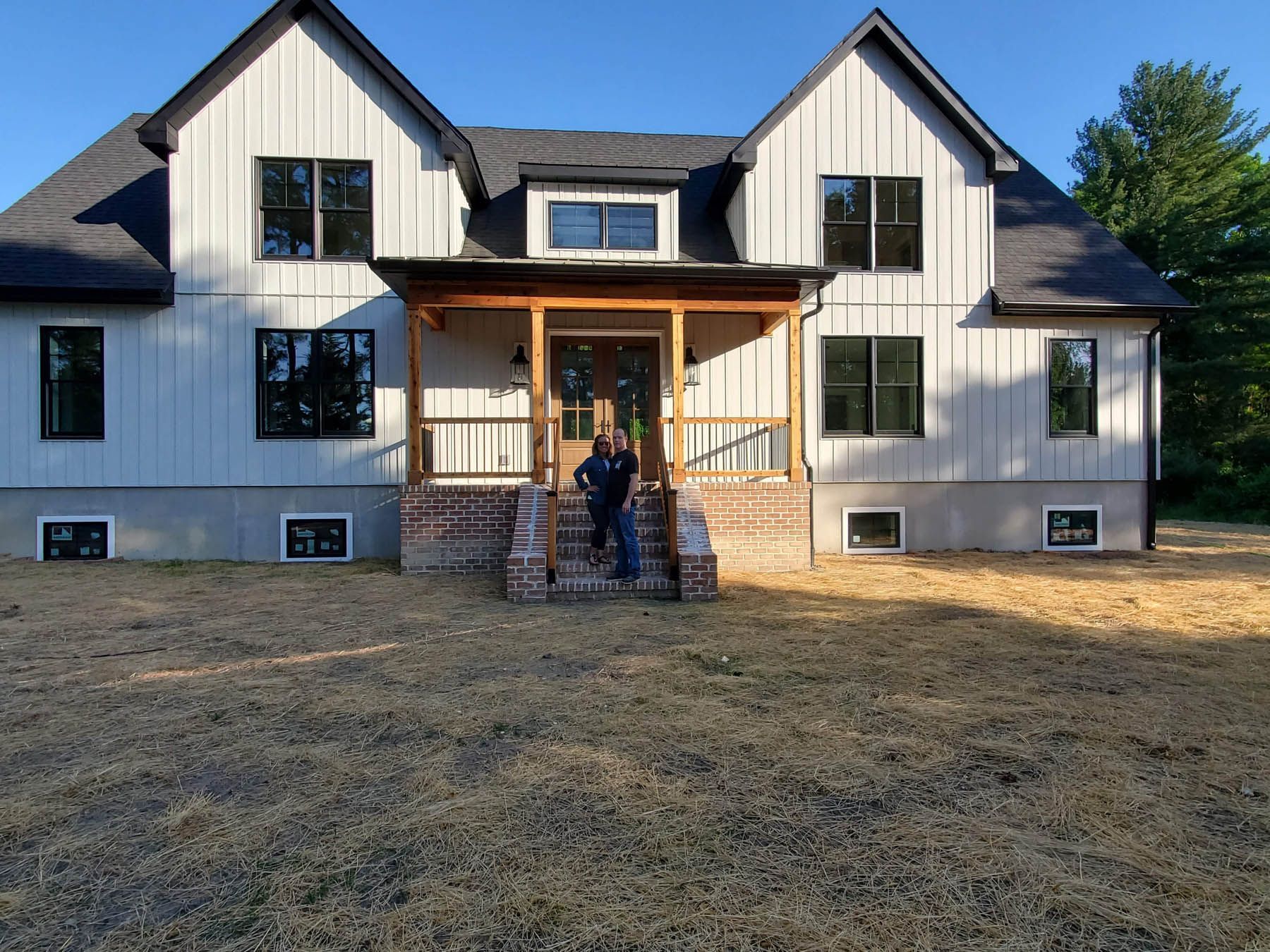 Two people stand on the steps of a new, light gray house with black windows and a wooden porch.