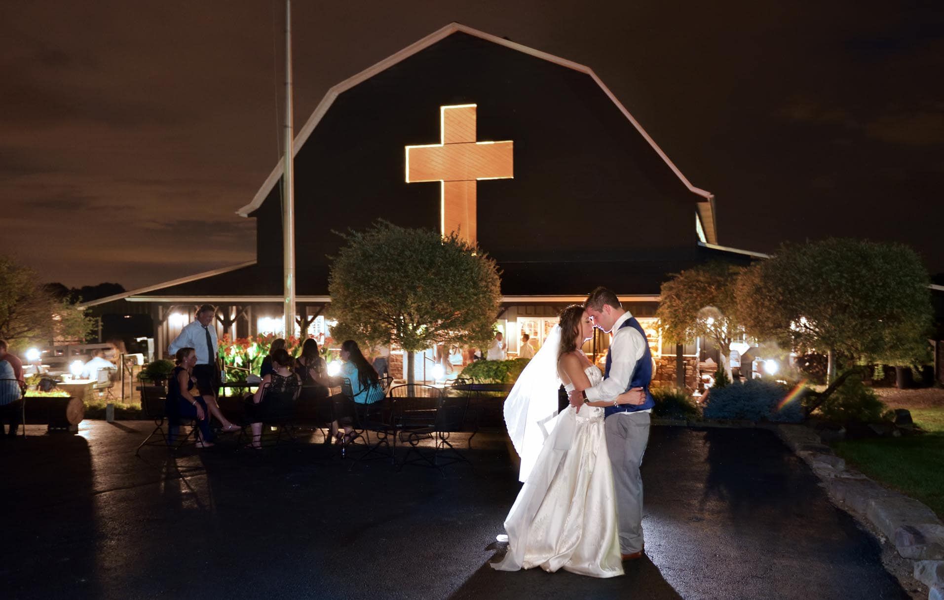 A bride and groom are kissing in front of a barn with a cross on it.