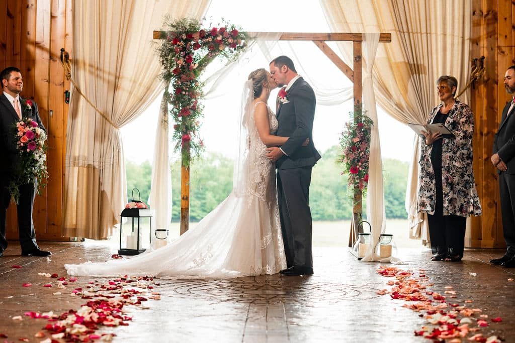 A bride and groom are kissing during their wedding ceremony in a barn.