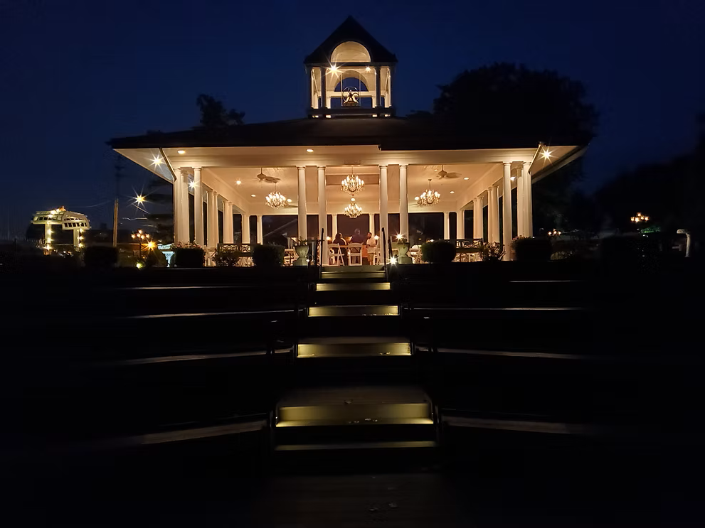 Illuminated gazebo at night, viewed from tiered steps. Lit columns, bell tower, and tables inside.
