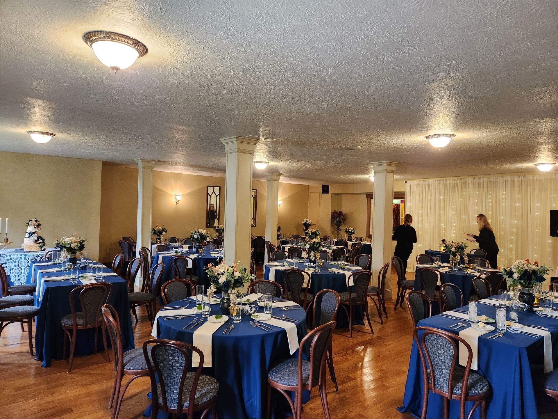 Formal event room with round tables draped in blue, decorated with flowers. Two people stand near a stage.