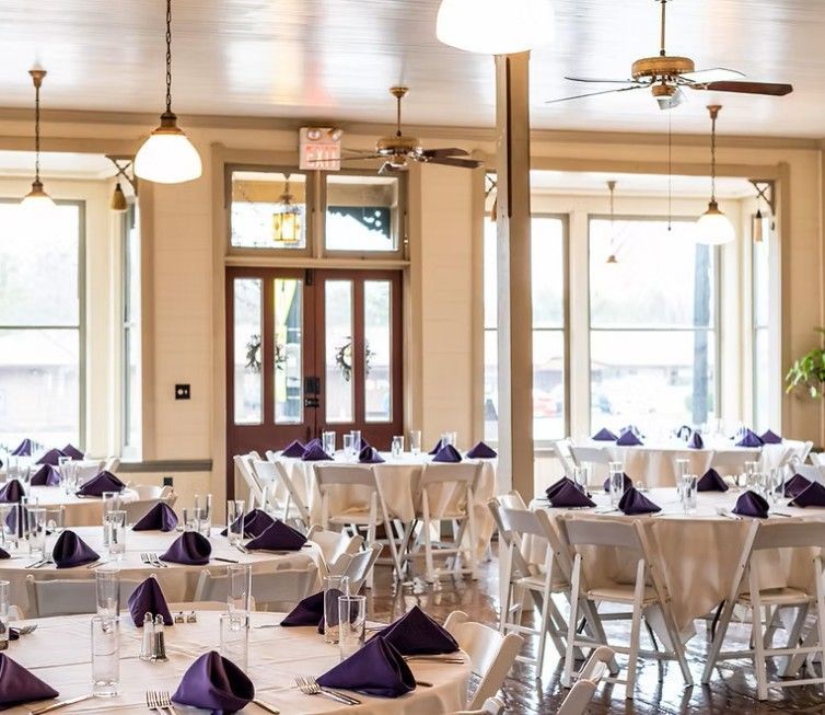 Elegant reception hall with round tables set for a formal event. White linens, purple napkins, and tall windows.