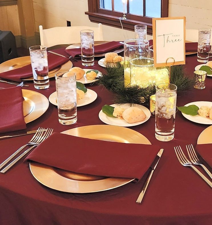 Round table set for a formal event, featuring gold chargers, burgundy napkins, and glasses.