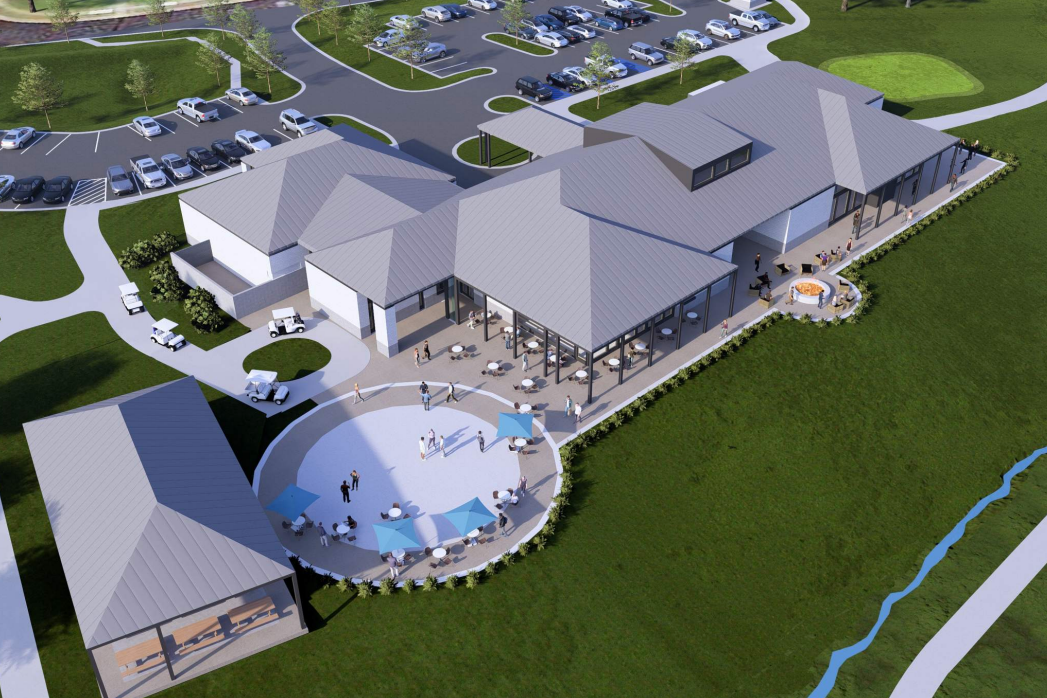 Aerial view of a modern building with a parking lot and a circular patio with umbrellas. Green grassy landscape surrounds.