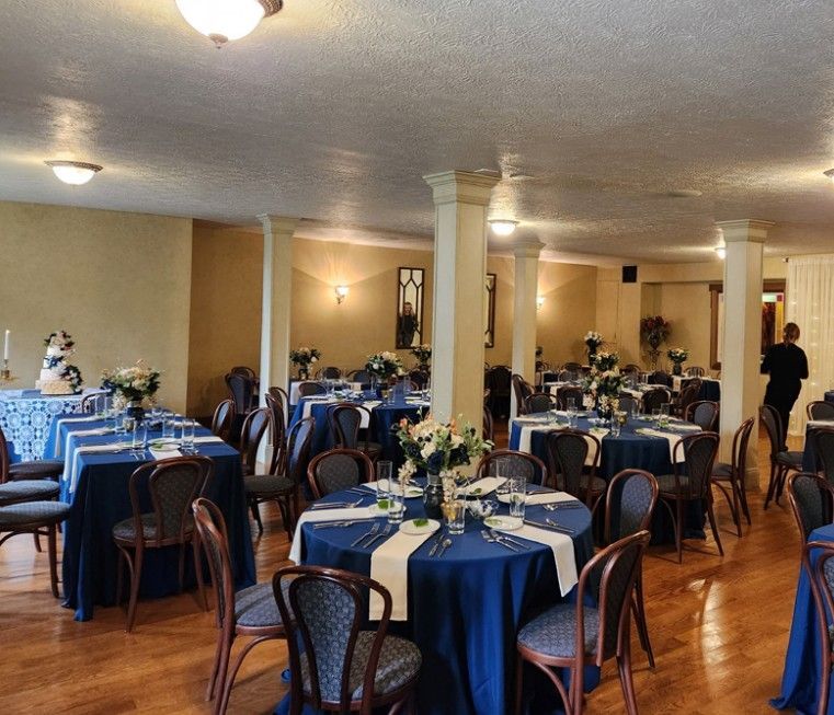 Reception hall with round tables set for a celebration, covered in blue tablecloths.