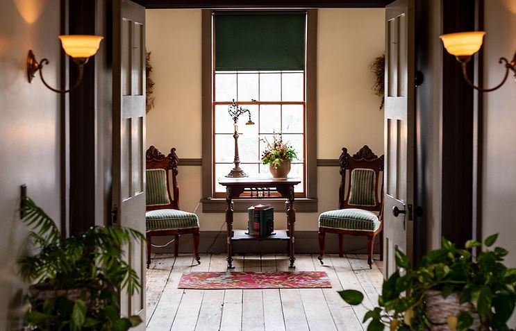 Open doorway to a room with chairs, table, window. Pale walls, dark wood trim. Two sconces.