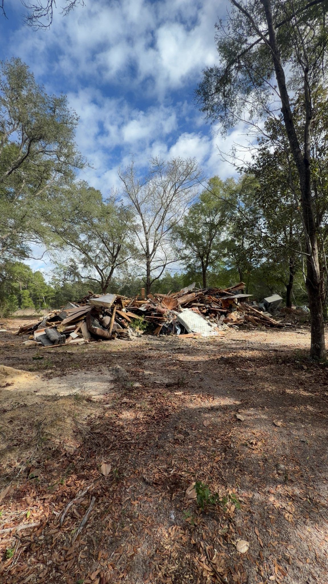 A large, scattered pile of construction debris and scrap metal sits in a clearing surrounded by trees under a blue sky.