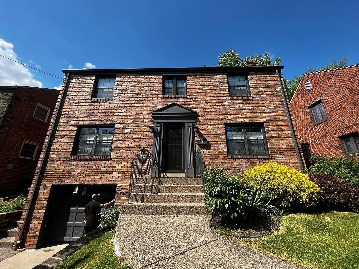Brick building with entrance steps, windows, and a green lawn. A garage door is visible on the left.