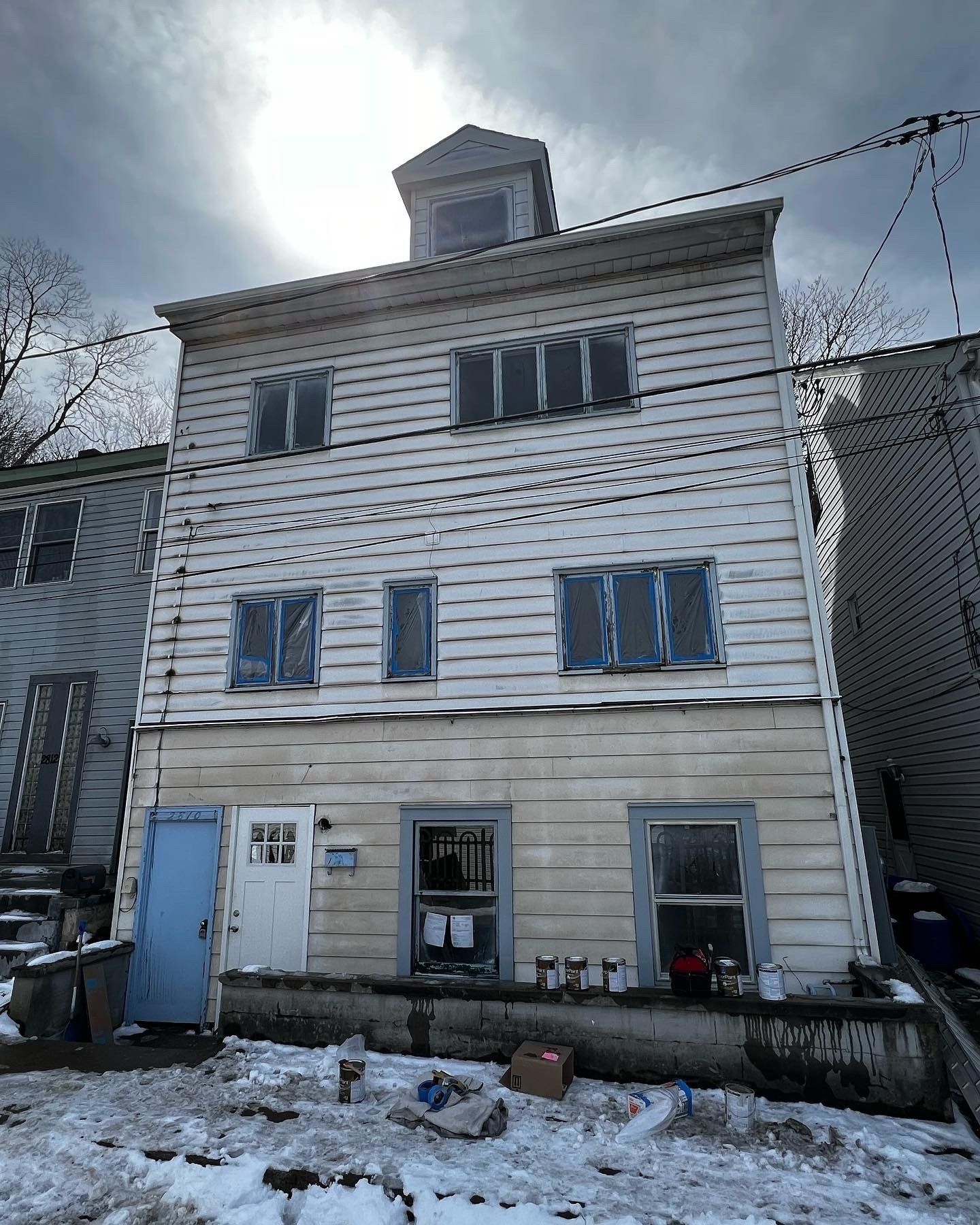 Three-story weathered house with boarded windows and a small tower. Snow and debris in front.