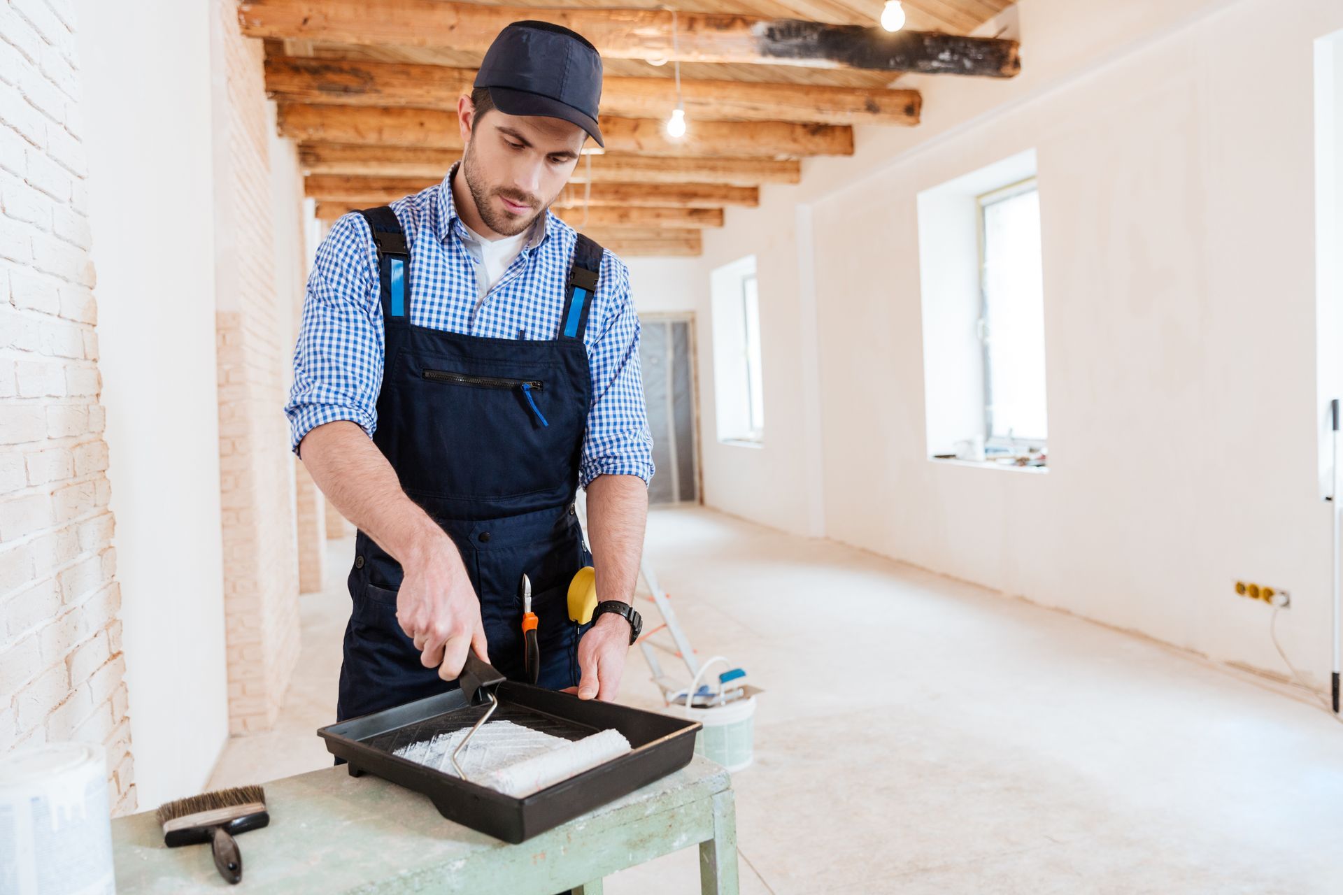 A painter in overalls loading a paint roller indoors.
