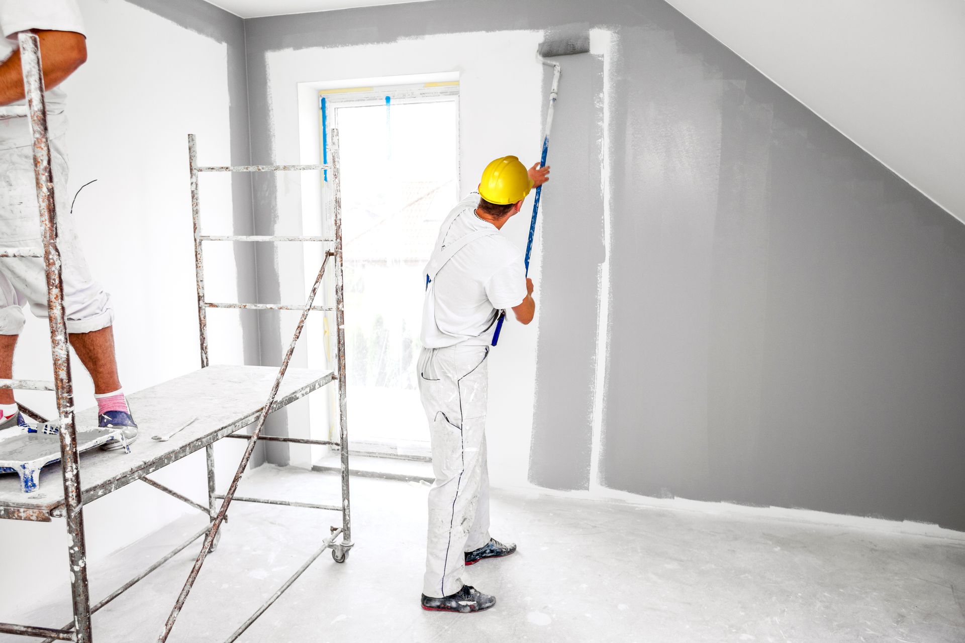 Painter in white coveralls rolling paint on gray interior wall.