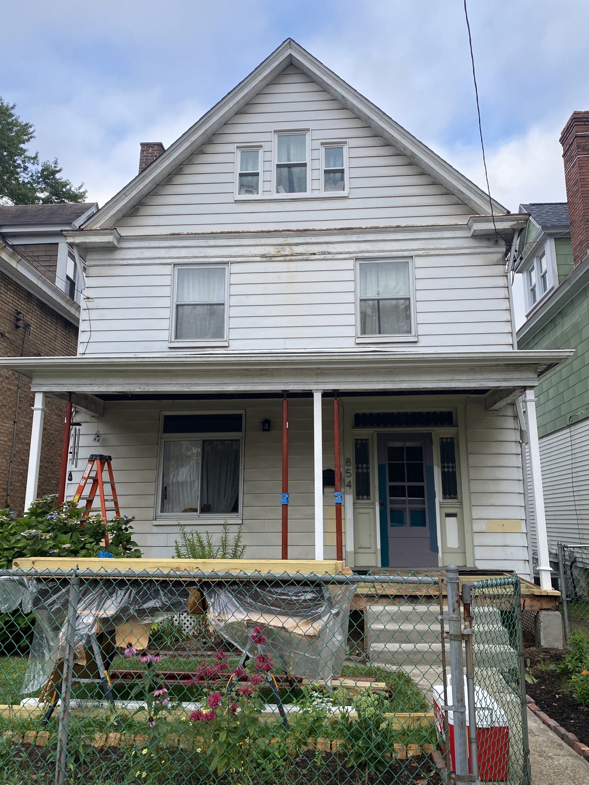 Two-story white house with porch and peeling paint. A fence and overgrown garden are in front.