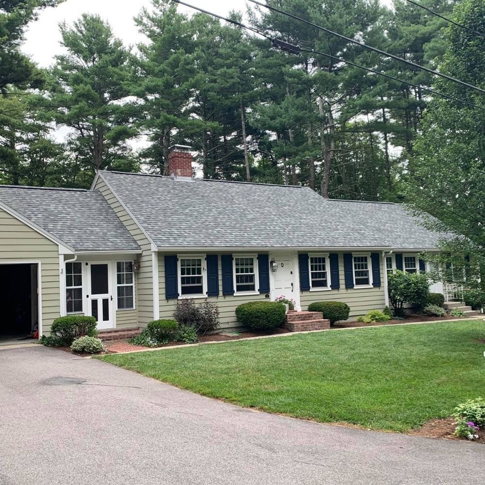 A single-story, light-green house with dark shutters and a gray roof, set against a backdrop of tall green trees.