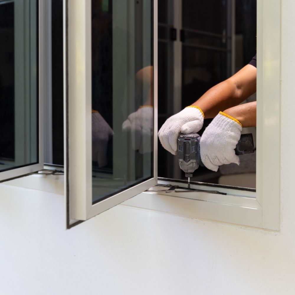 A person wearing white gloves uses a cordless drill to install or repair a white aluminum window frame.
