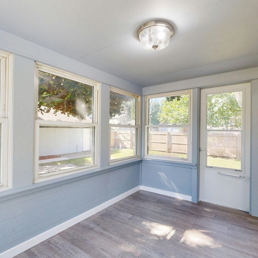 A sunroom with light blue walls, wood-look flooring, large windows, and a glass-pane door leading to a backyard.