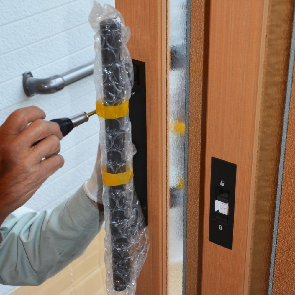 A person in a light-colored uniform uses a screwdriver to attach a wrapped handle to a wood-toned door frame.