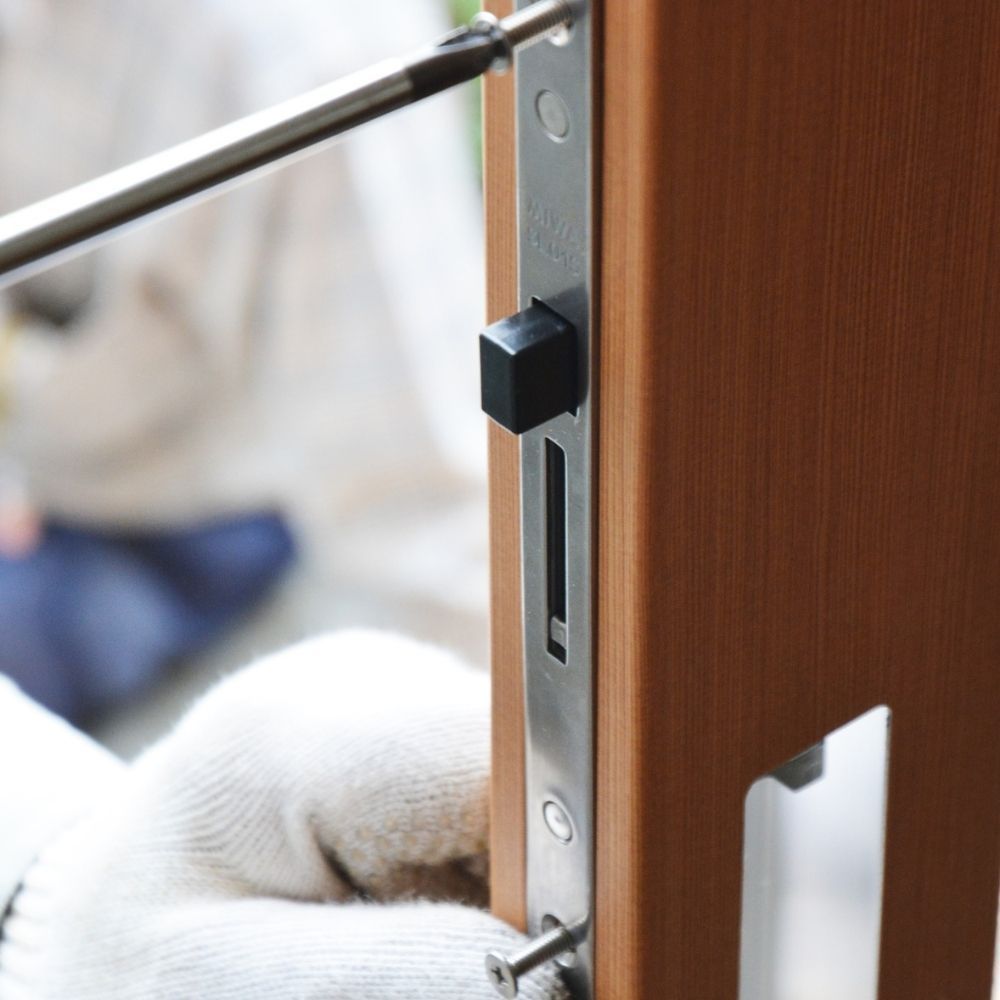 A gloved hand uses a screwdriver to install a metal mortise lock mechanism into the side of a wooden door.
