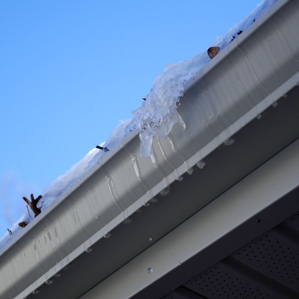 Melting snow and icicles clinging to the edge of a metal house gutter against a clear blue sky.