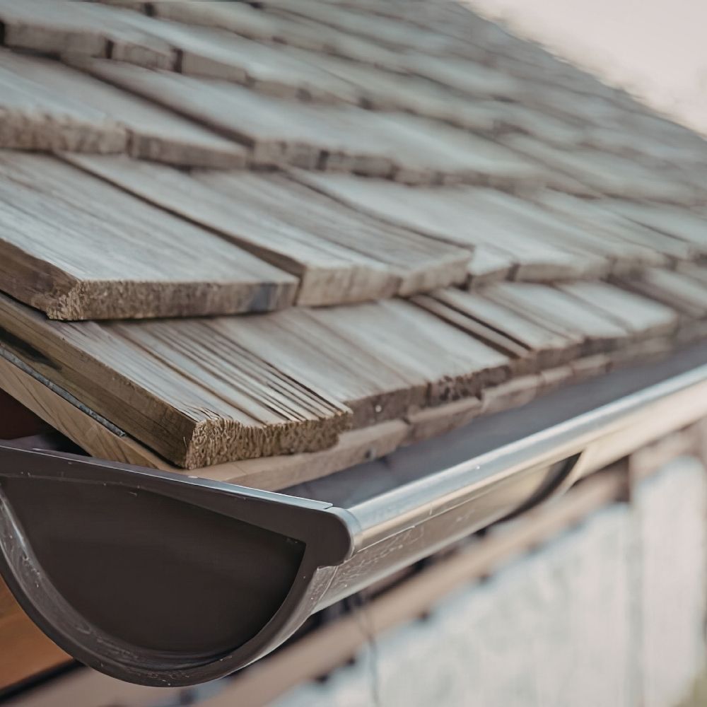 A dark brown half-round gutter mounted along the edge of a roof with weathered wood shingles.