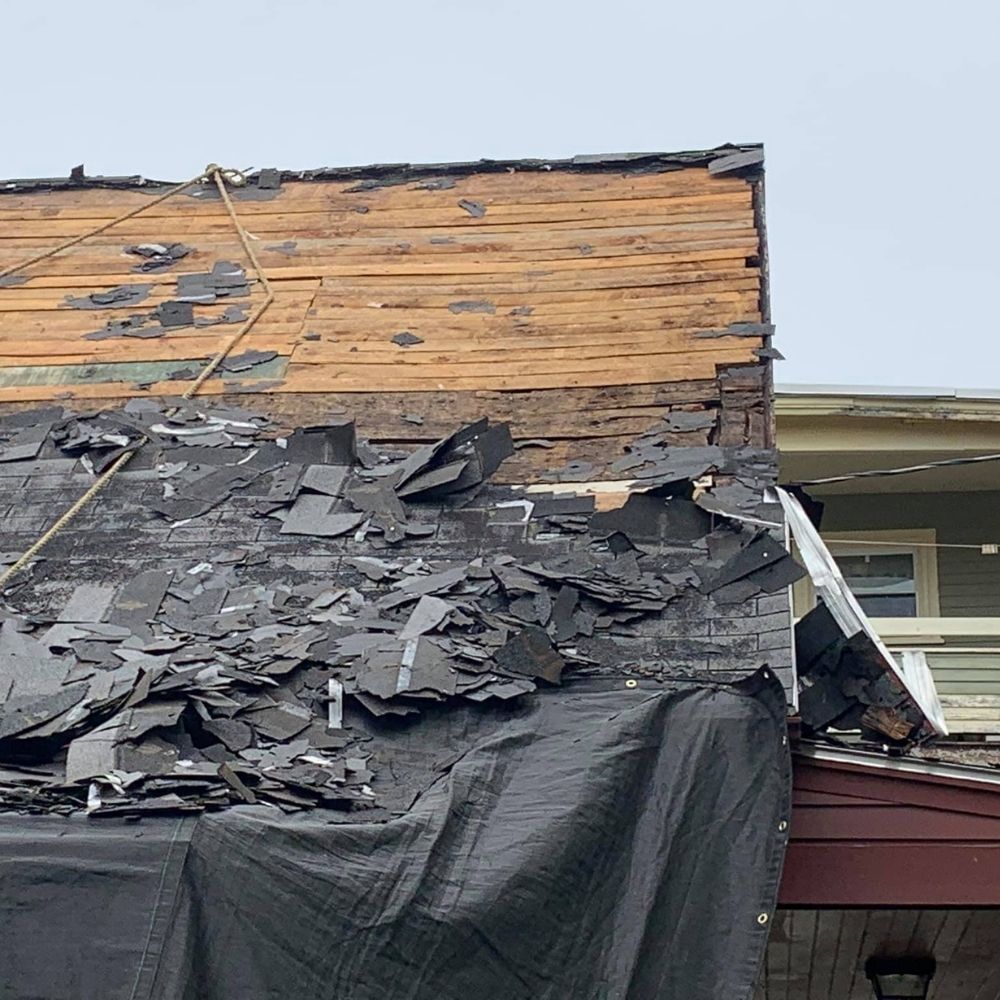 A house with a roof undergoing repairs, showing exposed wooden planks and piles of torn-off black shingles.