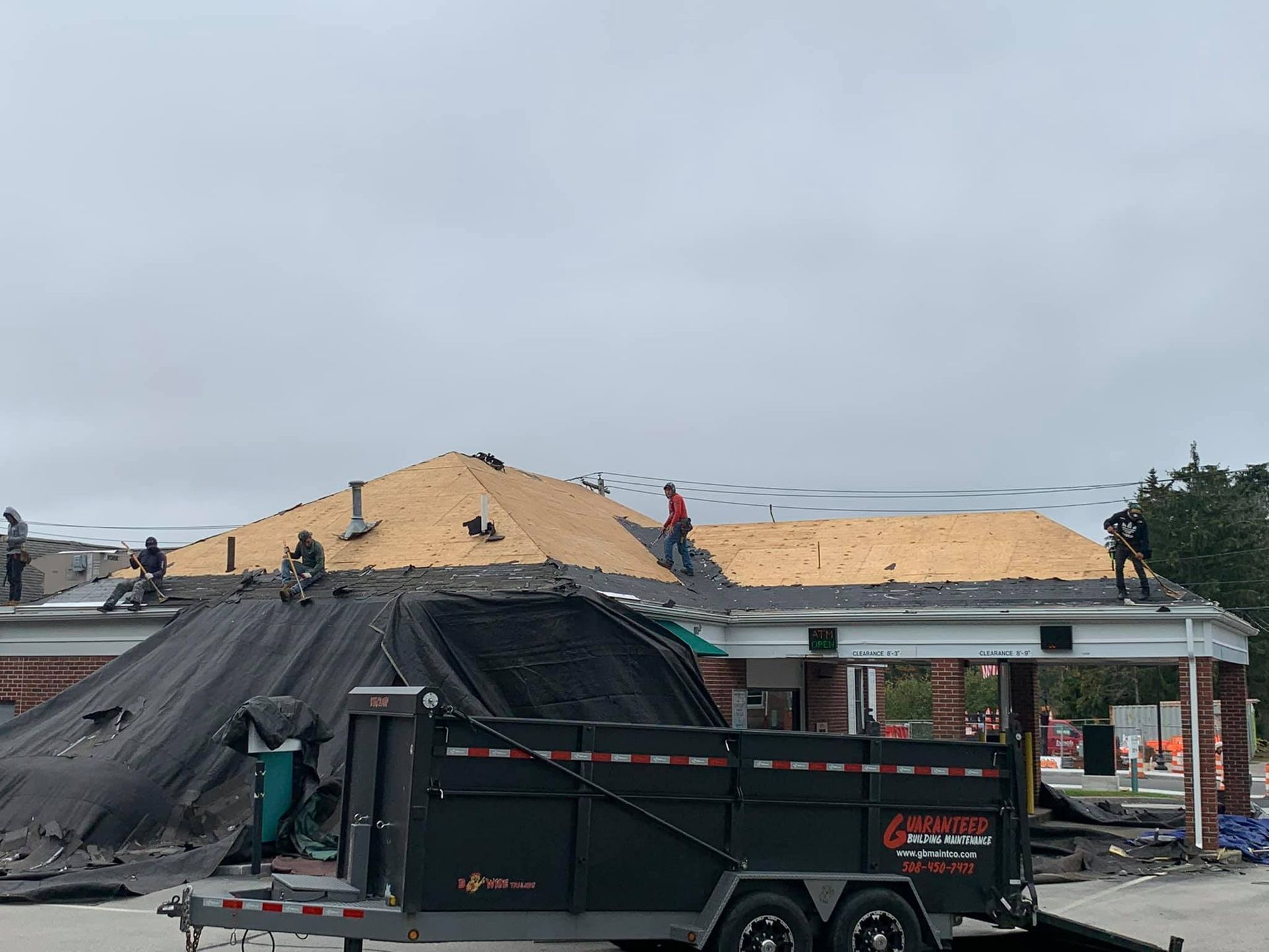 Roofers working on a building with exposed plywood decking, with a large black debris trailer parked in front.
