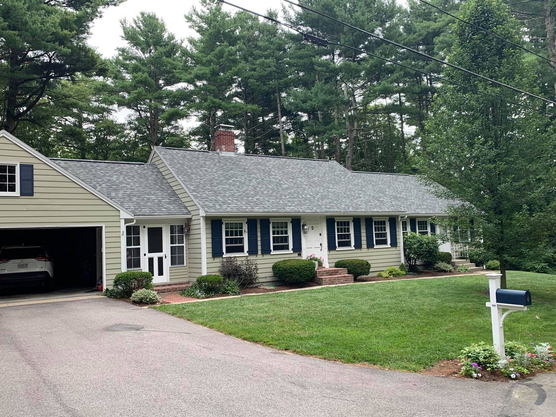 A one-story suburban ranch house with light siding, dark blue shutters, a garage, and a manicured lawn among tall trees.