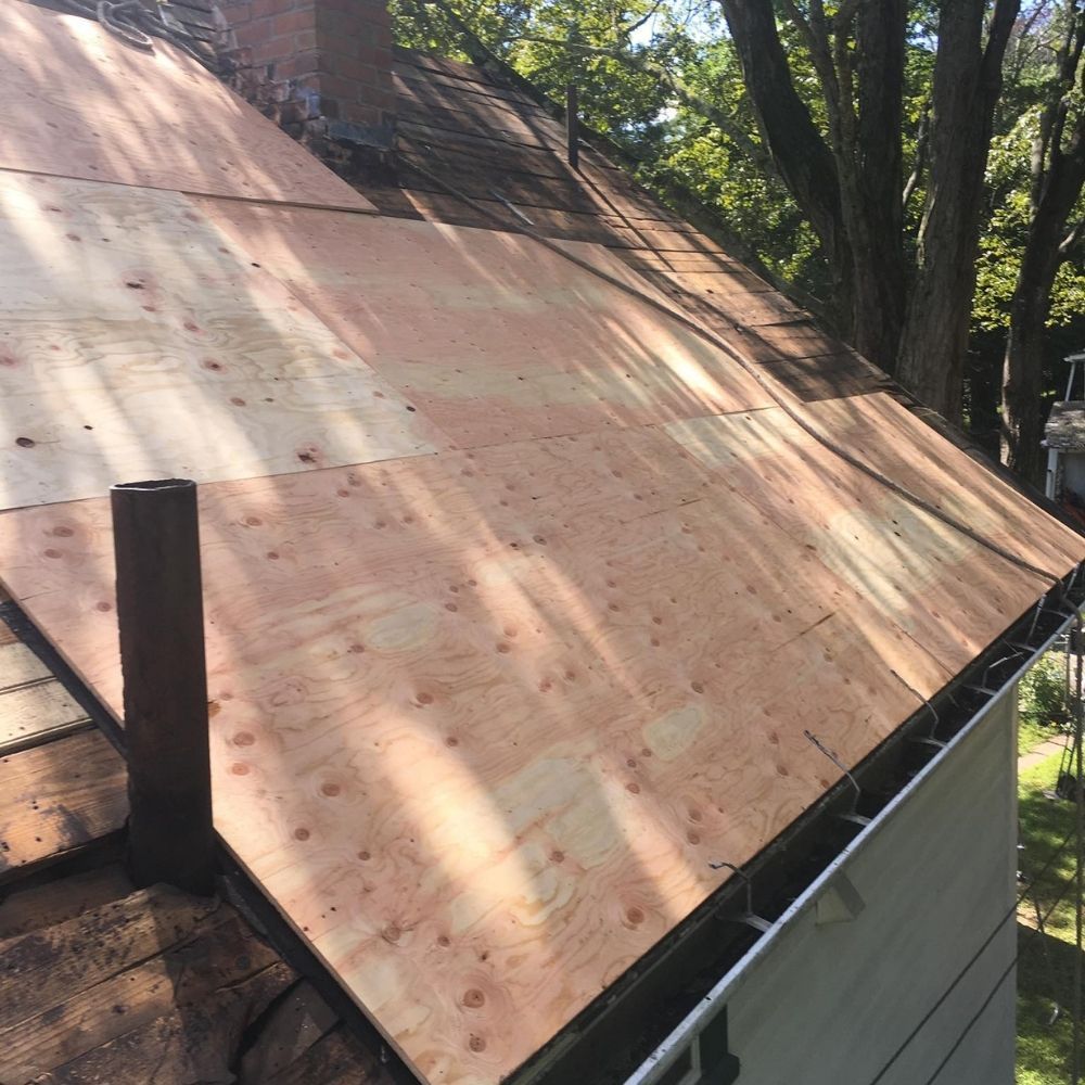 A roof under renovation, featuring new plywood sheathing laid across the rafters, with a chimney and plumbing vent visible.