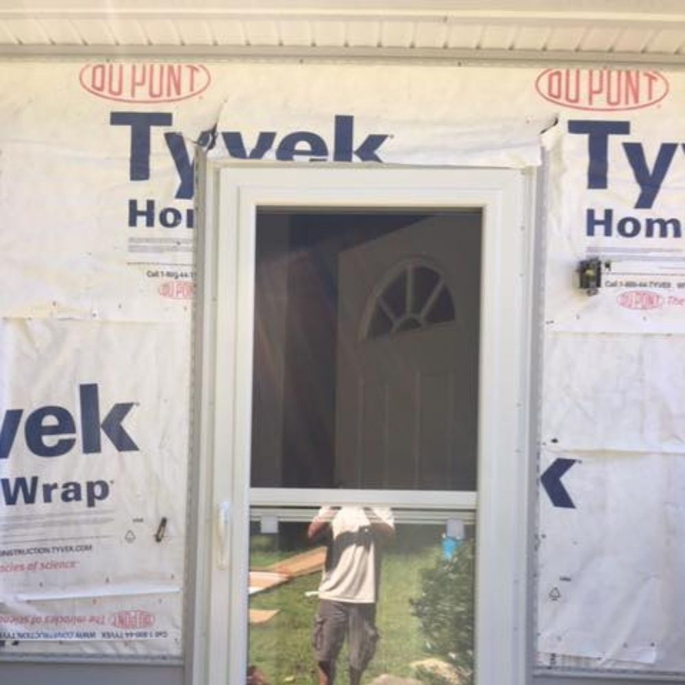 A screen door installed on a house frame covered in Tyvek wrap, with a person reflected in the glass.