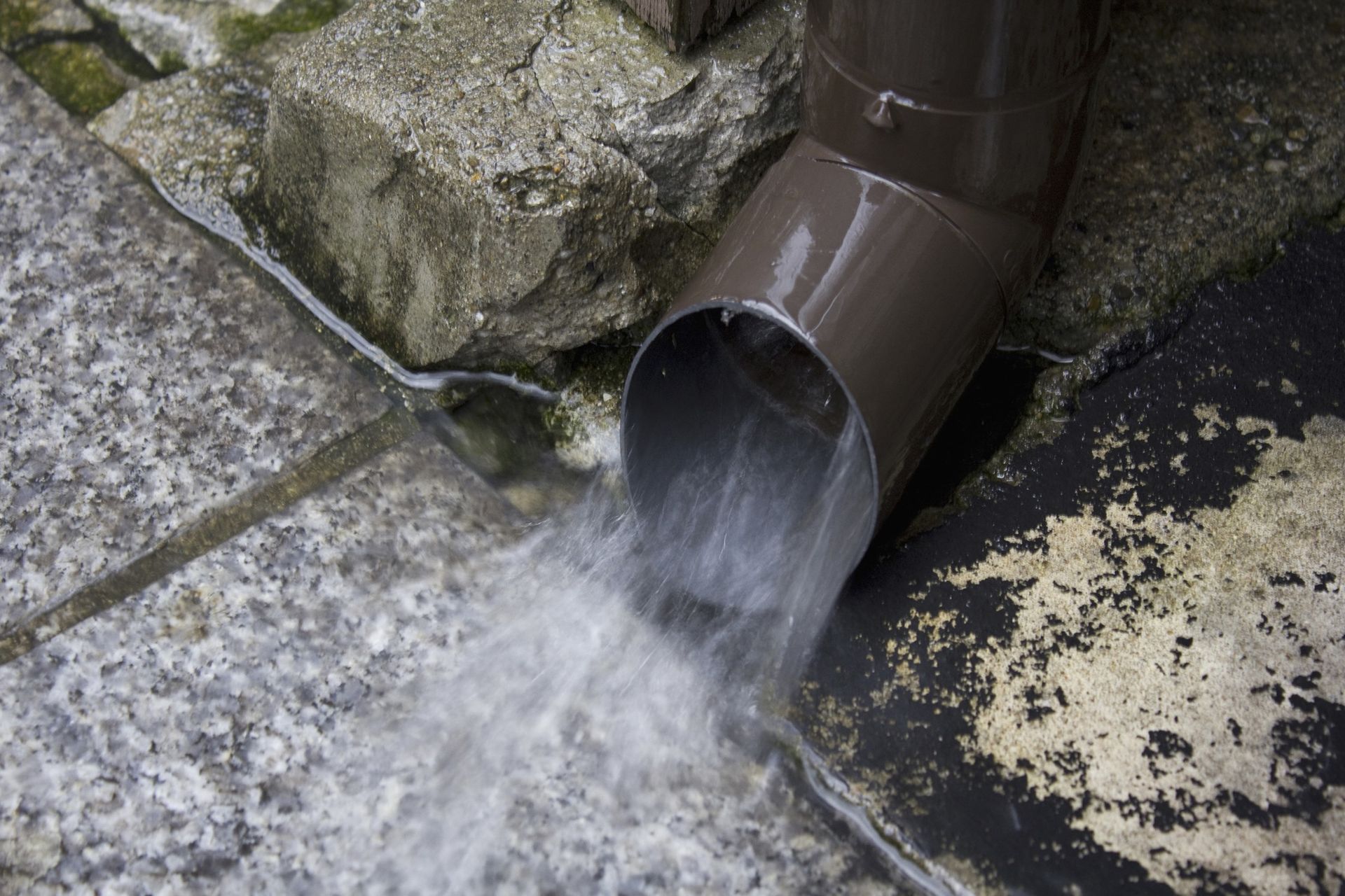 Water flows from a brown downspout onto a stone ground surface.