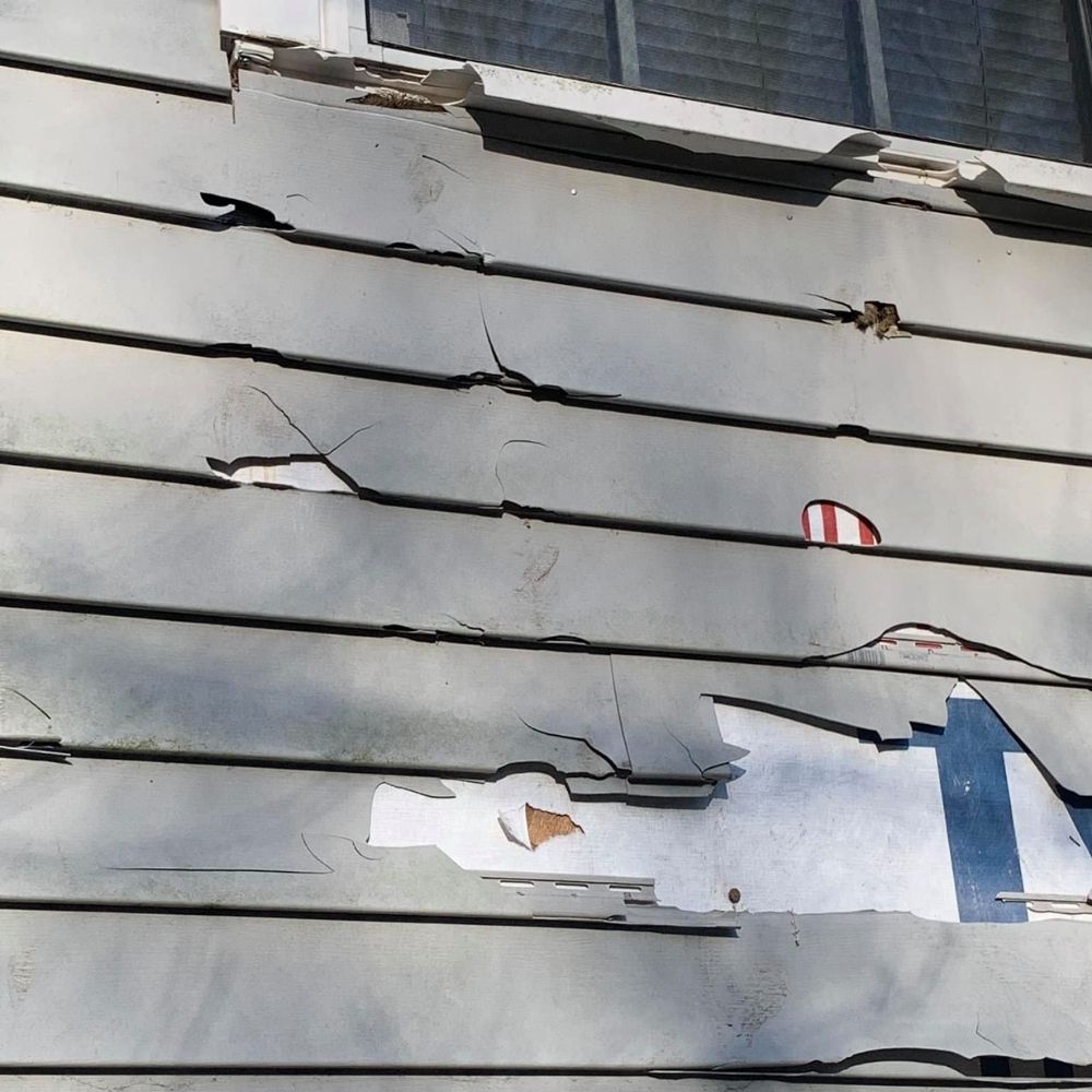 Close-up of damaged, gray horizontal siding on the exterior of a home, showing cracks and missing sections.