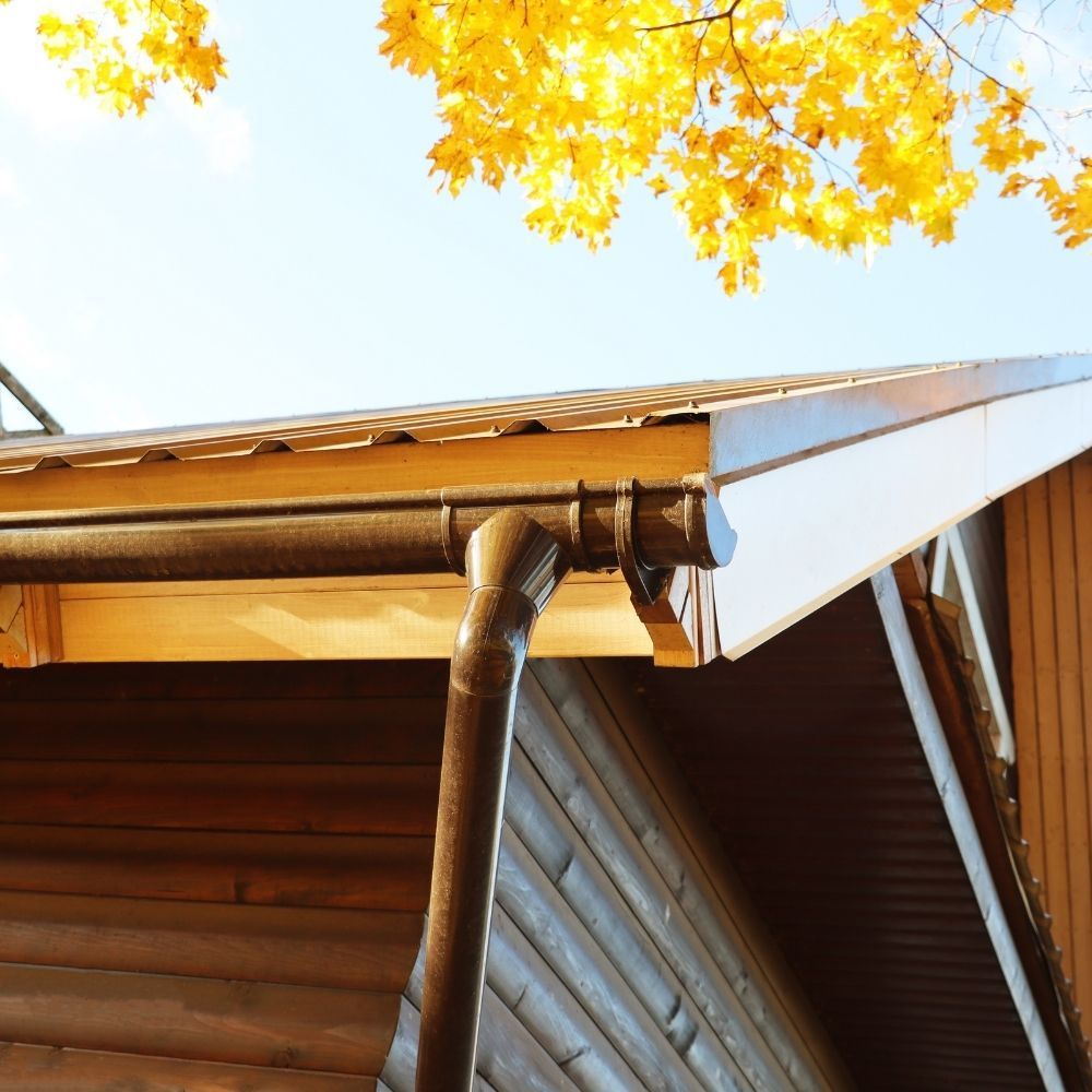 A brown metal gutter and downspout attached to the wooden eaves of a cabin under autumn foliage against a blue sky.