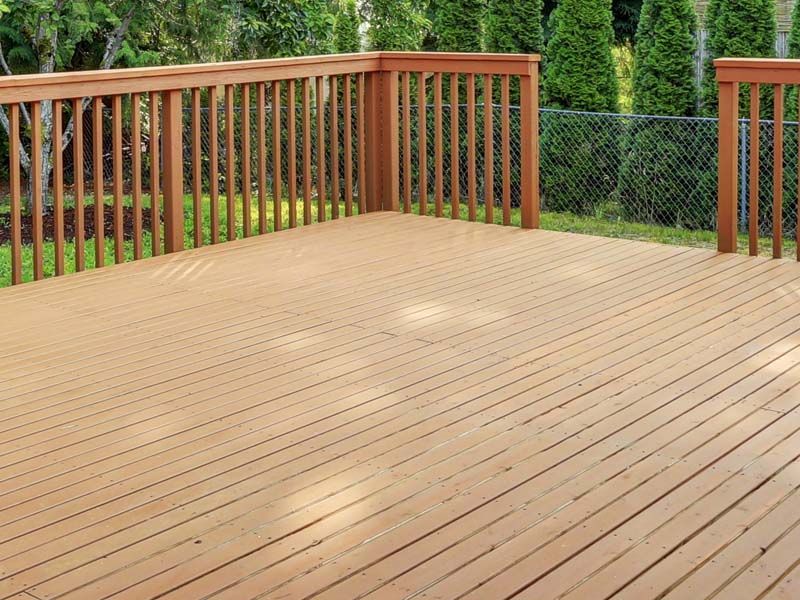 A wooden backyard deck with a railing, overlooking a lawn with green trees and a chain-link fence.