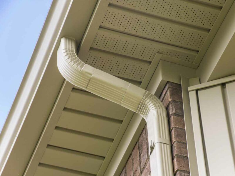 A tan-colored metal downspout curves from the roof gutter down the side of a brick wall against a light-colored soffit.