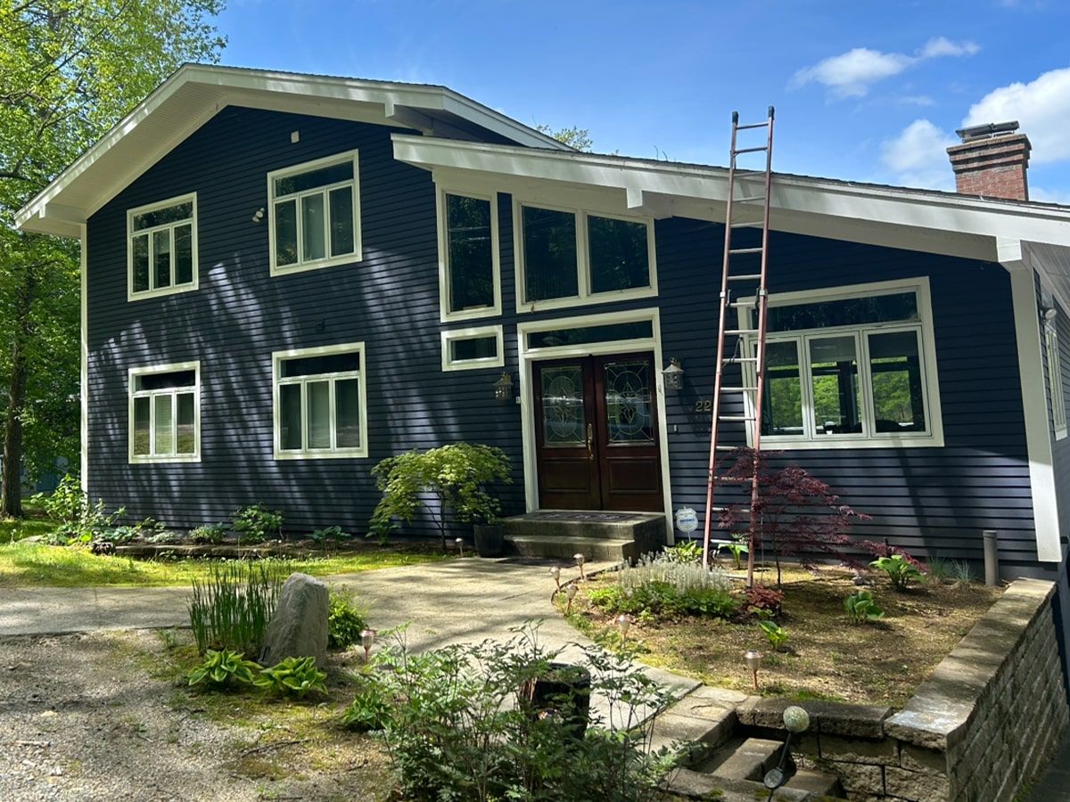 A two-story dark blue house with white trim, large windows, a double front door, and a tall ladder leaning on the roof.