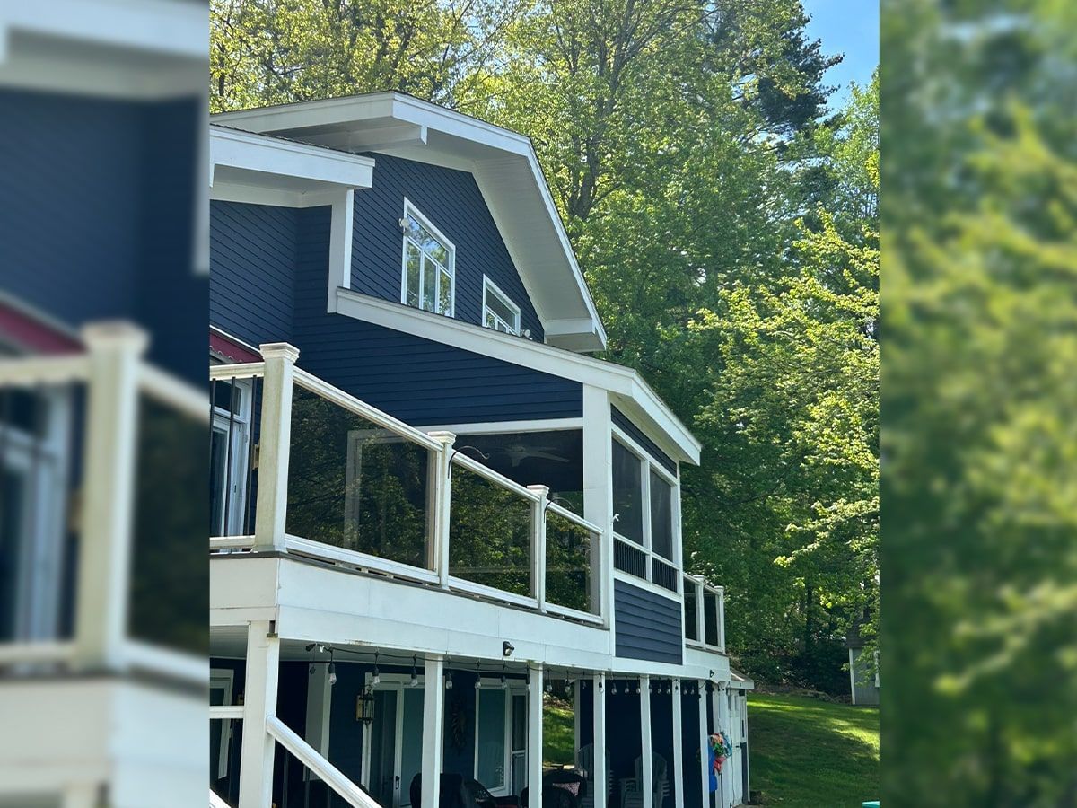 A two-story blue house with a white wraparound deck and glass railings, surrounded by lush green trees on a sunny day.