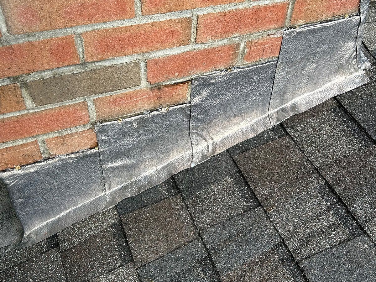 Lead flashing installed along the base of a brick chimney where it meets the asphalt shingle roof.